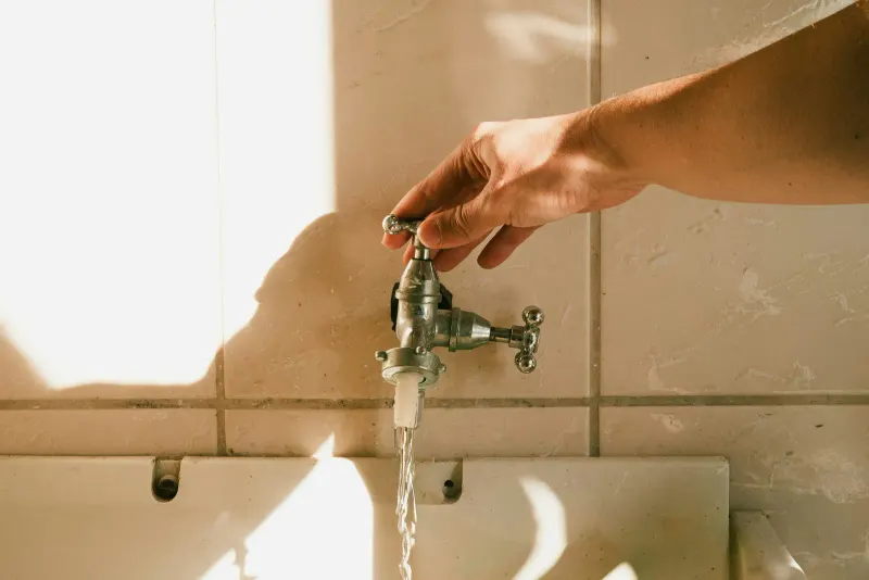 A hand turning on a wall tap in warm morning light, a thin stream of water running from it — the same supply most cities disinfect with chlorine or chloramine