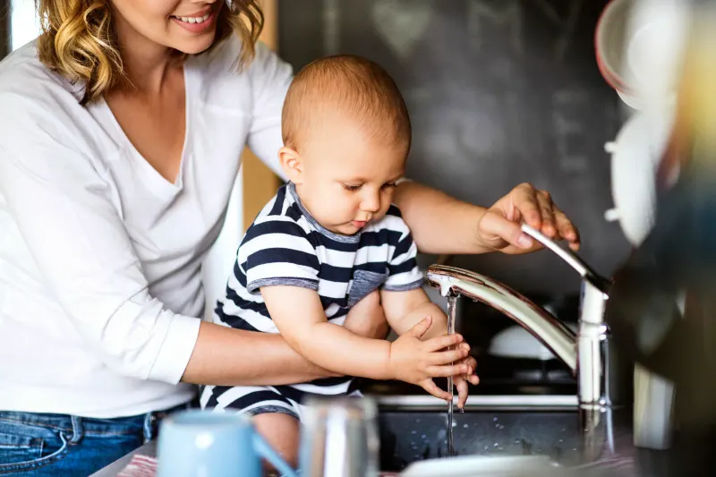Mother helping baby reach toward a kitchen faucet with running water