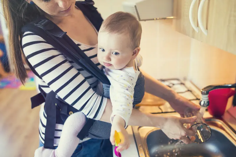 Mother washing up at the kitchen sink with her baby in a carrier — on a private well, this tap is also the water for the bottle