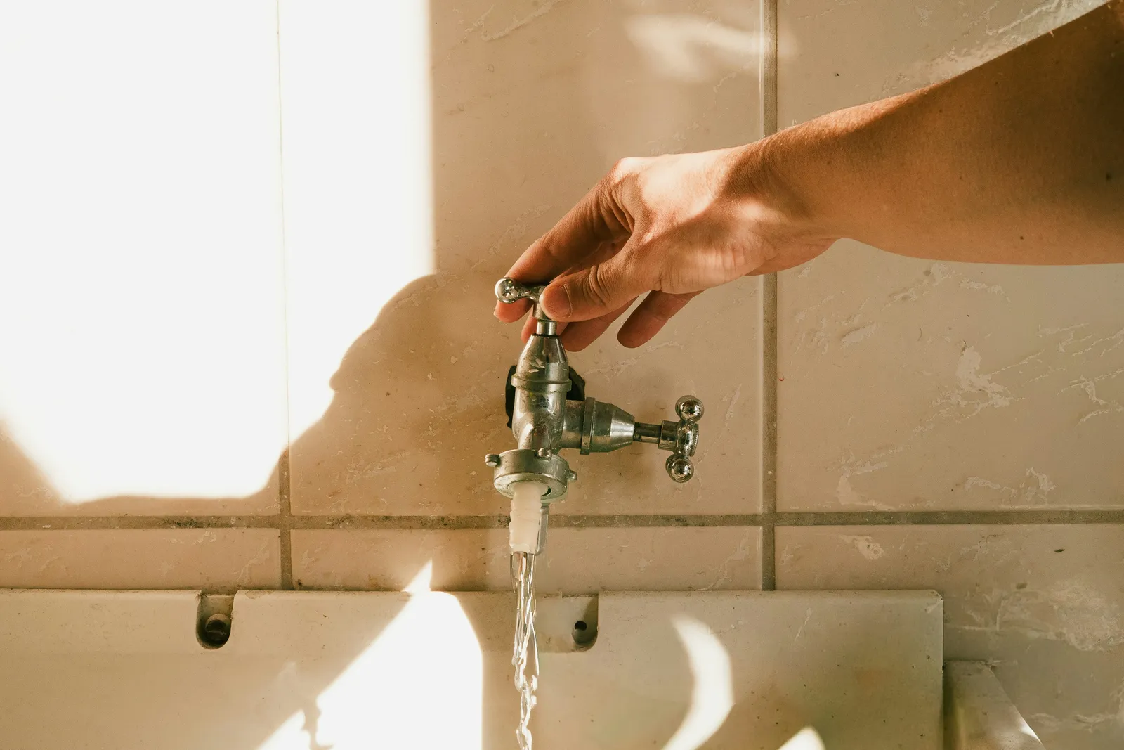A hand turning on a wall tap in warm morning light, a thin stream of water running from it — the same supply most cities disinfect with chlorine or chloramine