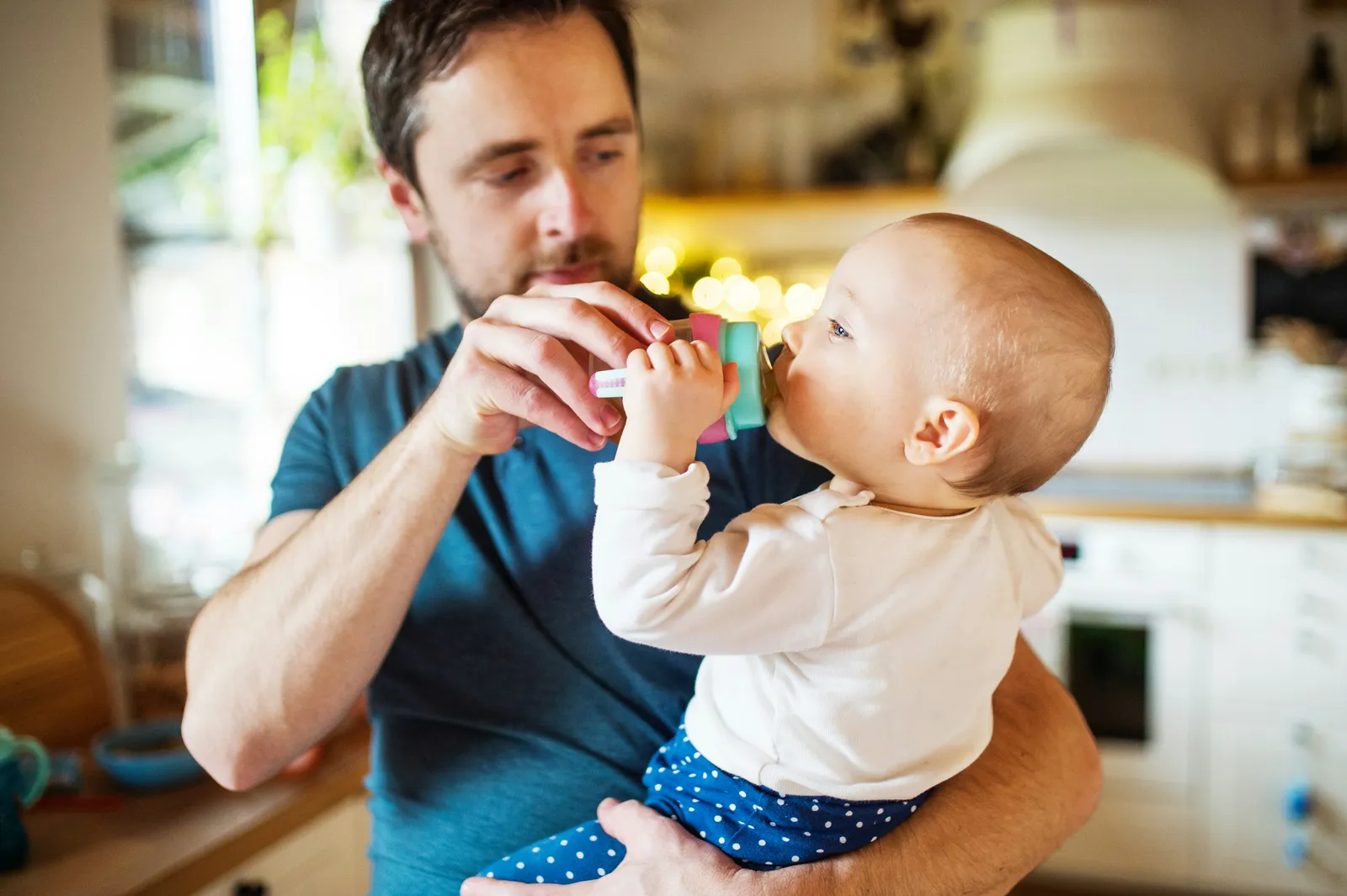 Father holding his baby daughter in a sunny kitchen while she drinks water from a small bottle
