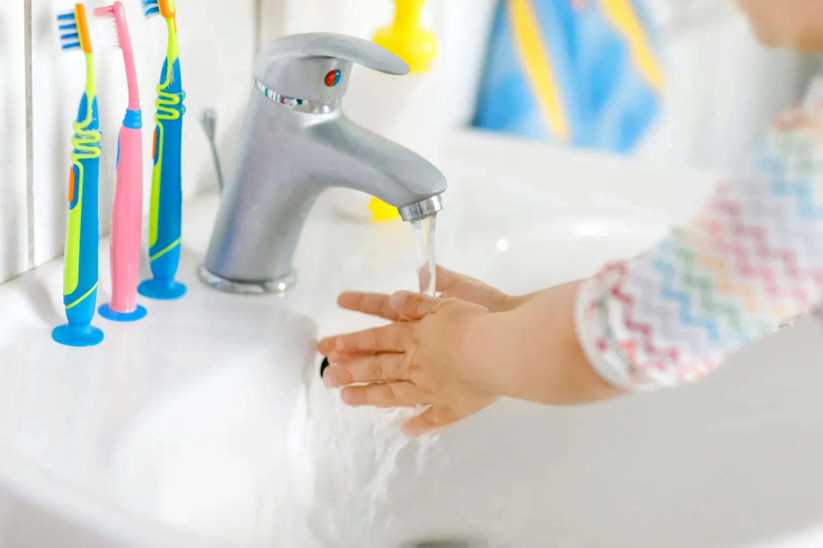 Child washing hands under a bathroom tap beside a cup of toothbrushes