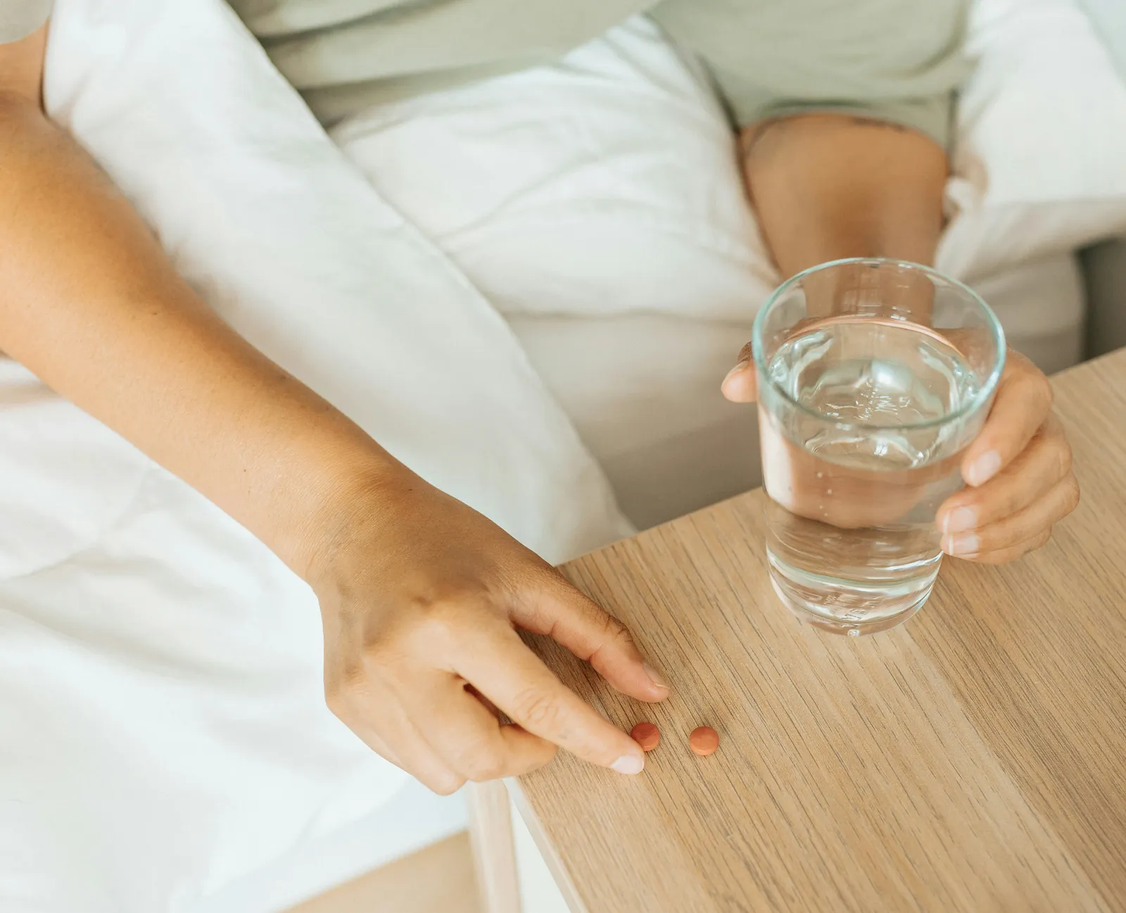 Hands reaching for a glass of water on a wooden bedside table in soft morning light