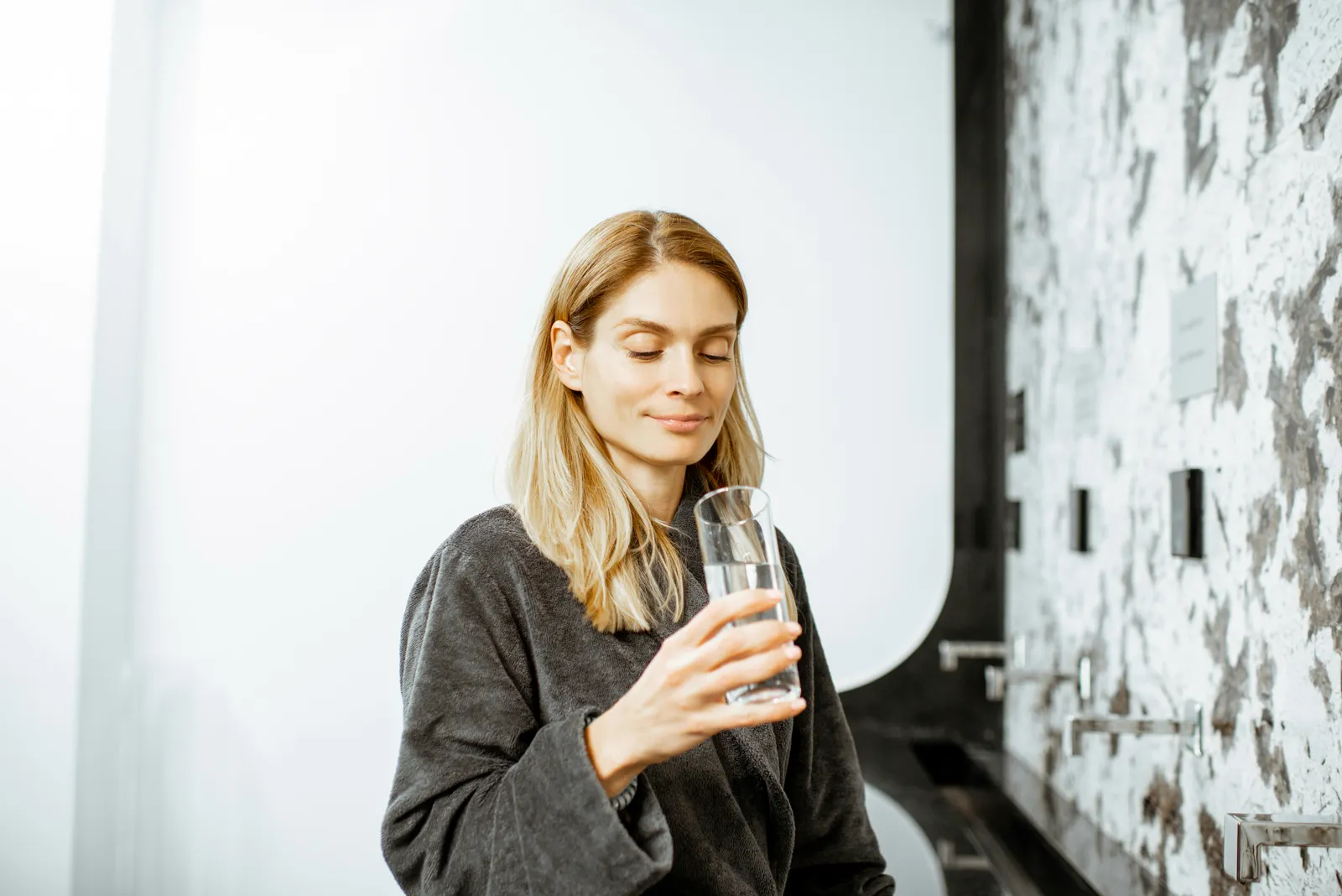 Woman in a robe calmly holding a clear glass of water — making an informed choice about drinking water quality