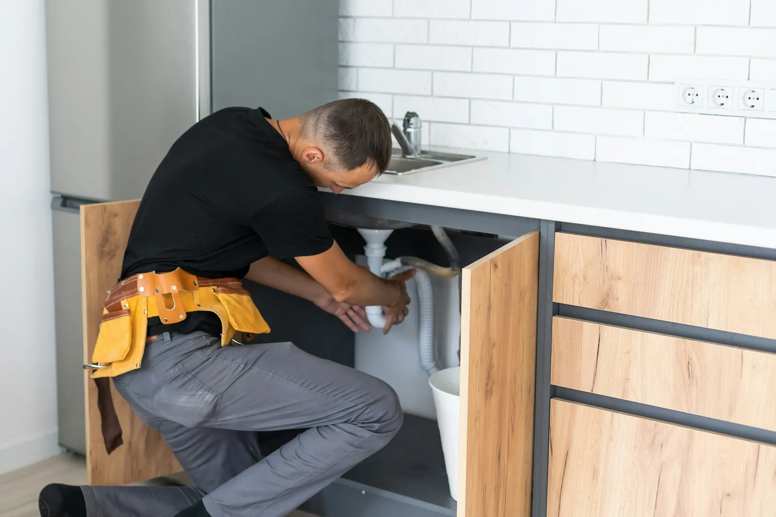 Installer kneeling at an open kitchen sink cabinet, fitting under-sink plumbing