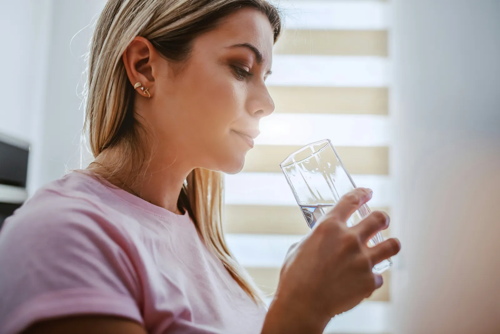 Woman drinking a glass of water by a bright kitchen window, the everyday choice between bottled spring water and filtered tap water