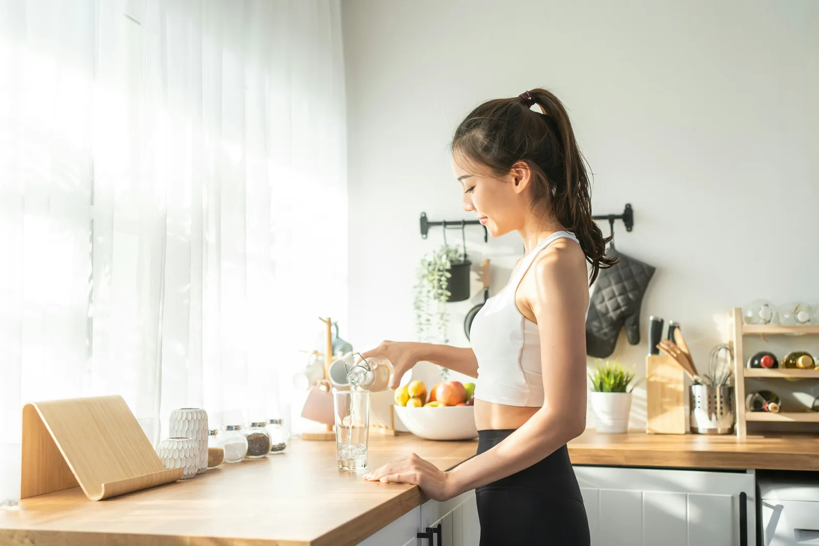 Woman pouring a glass of water at a sunlit kitchen counter, the everyday moment where distilled versus reverse osmosis water becomes a real choice