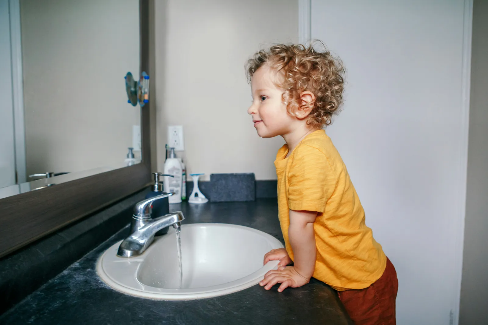 Curly-haired toddler leaning over a bathroom sink to watch the running tap
