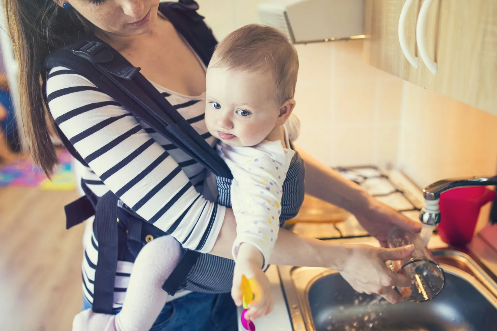 Mother washing up at the kitchen sink with her baby in a carrier — on a private well, this tap is also the water for the bottle