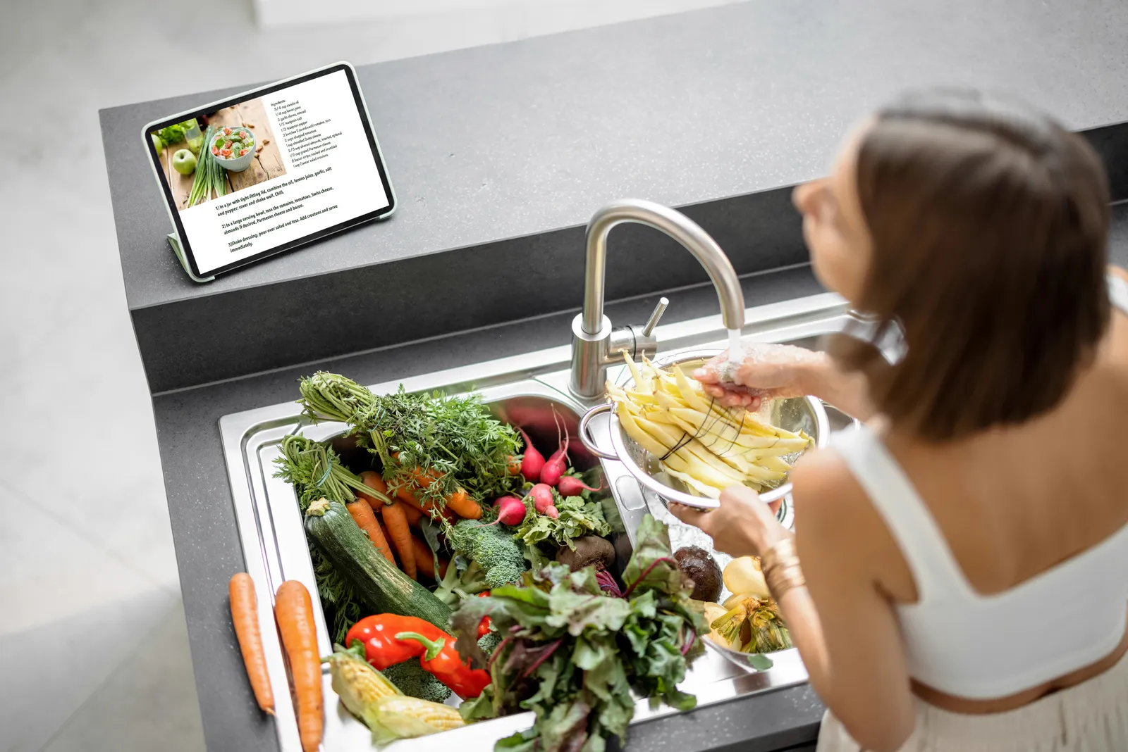 Woman rinsing vegetables at a modern kitchen sink with a tablet propped nearby — clean water at the kitchen tap is what most people want to protect first